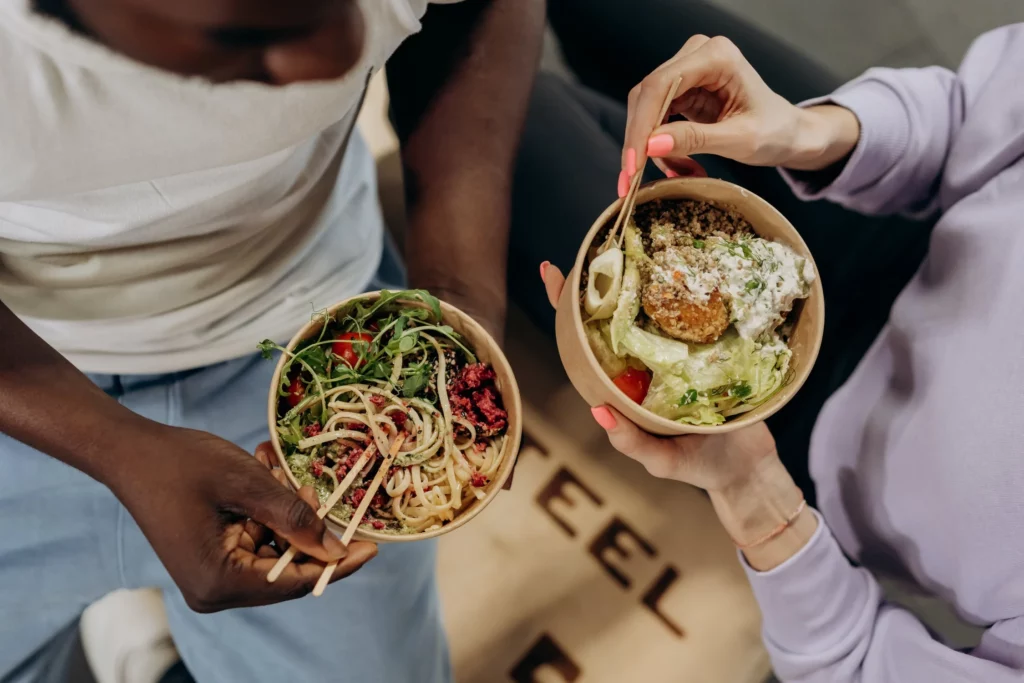 Two women holding bowls of healthy food and eating with chopsticks