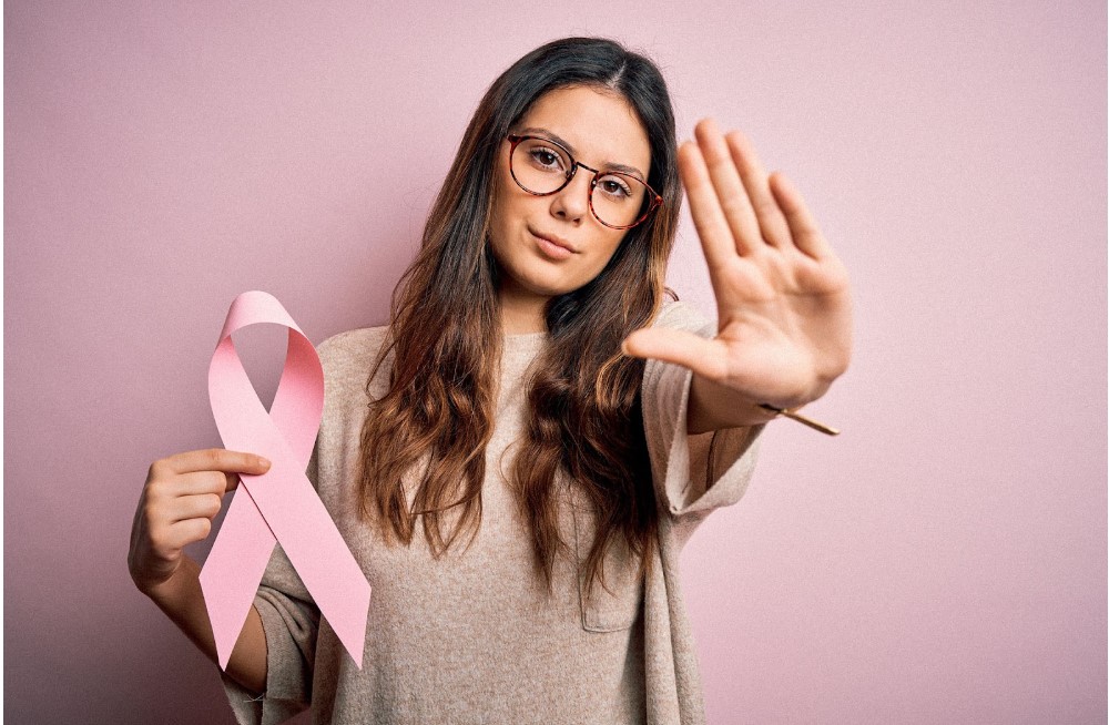 A young brunette woman holding the pink breast cancer ribbon in one hand, and holding up the other to signal a halt to breast cancer.