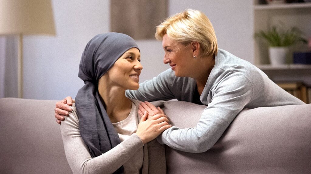 Une femme souriante atteinte d'un cancer, portant un foulard sur la tête, réconfortée par une proche.