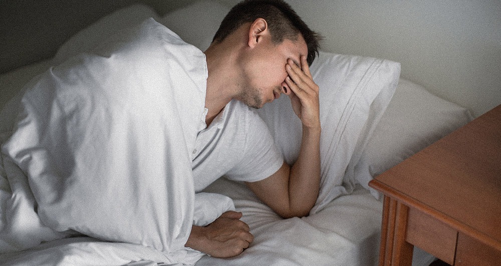 A man holding his head after a bad nights sleep struggling to get out of bed.