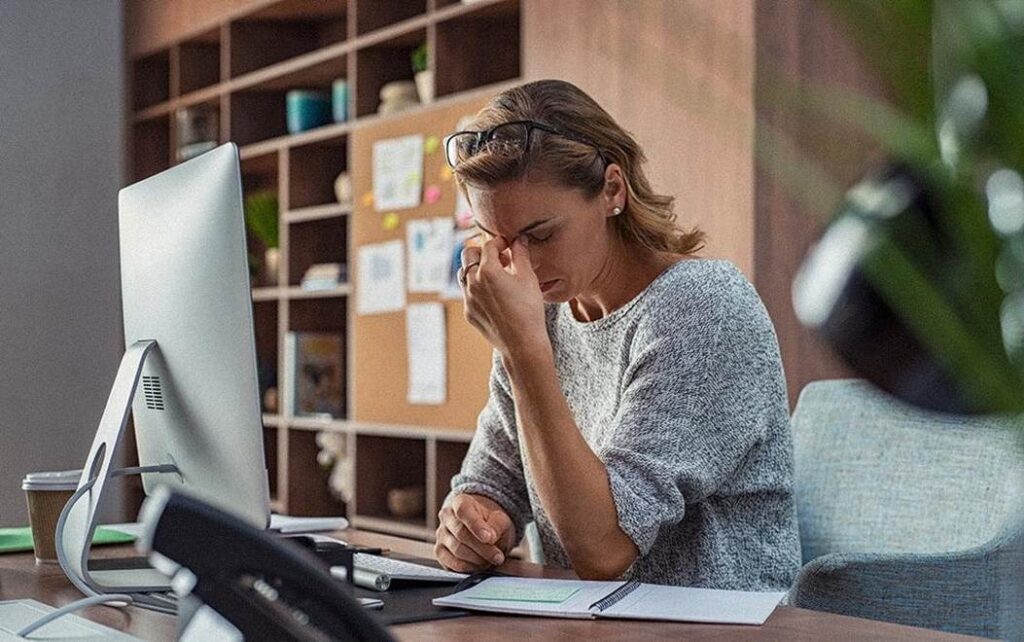 Une femme d'affaires à son bureau devant un ordinateur, dans un lieu de travail. Elle se tient la tête pour signifier un mal de tête, de la fatigue et du stress.
