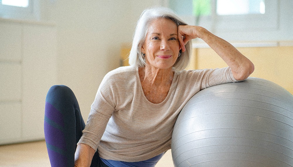 Smiling elderly woman resting on a swiss ball at home.