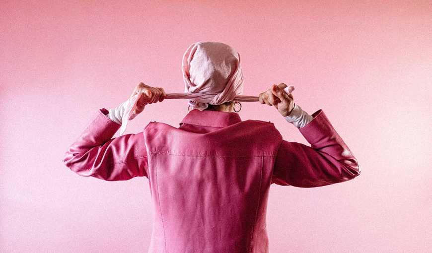 Une femme, sur fond rose, nouant un foulard rose autour de sa tête pour lutter contre le cancer du sein.