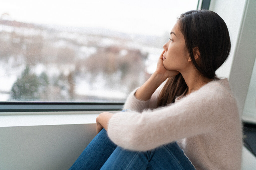 Femme assise près d’une fenêtre, regardant un paysage hivernal enneigé.
