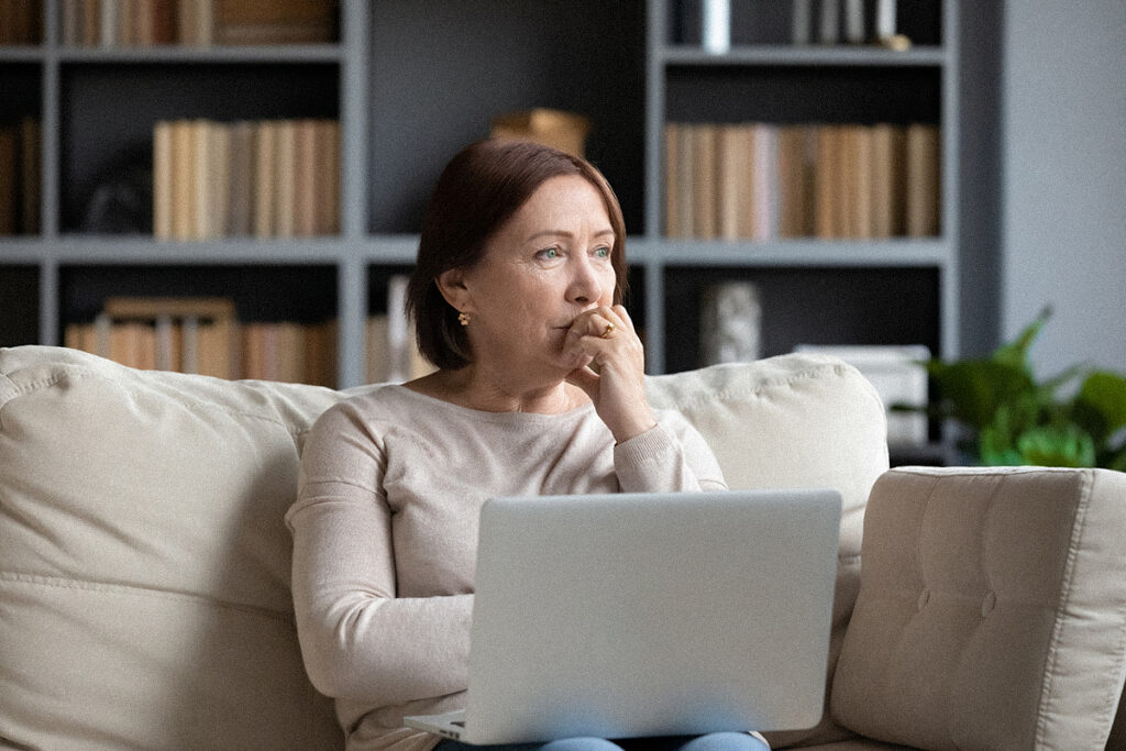 Middle-aged woman with laptop sitting indoors looking thoughtfully out a window.