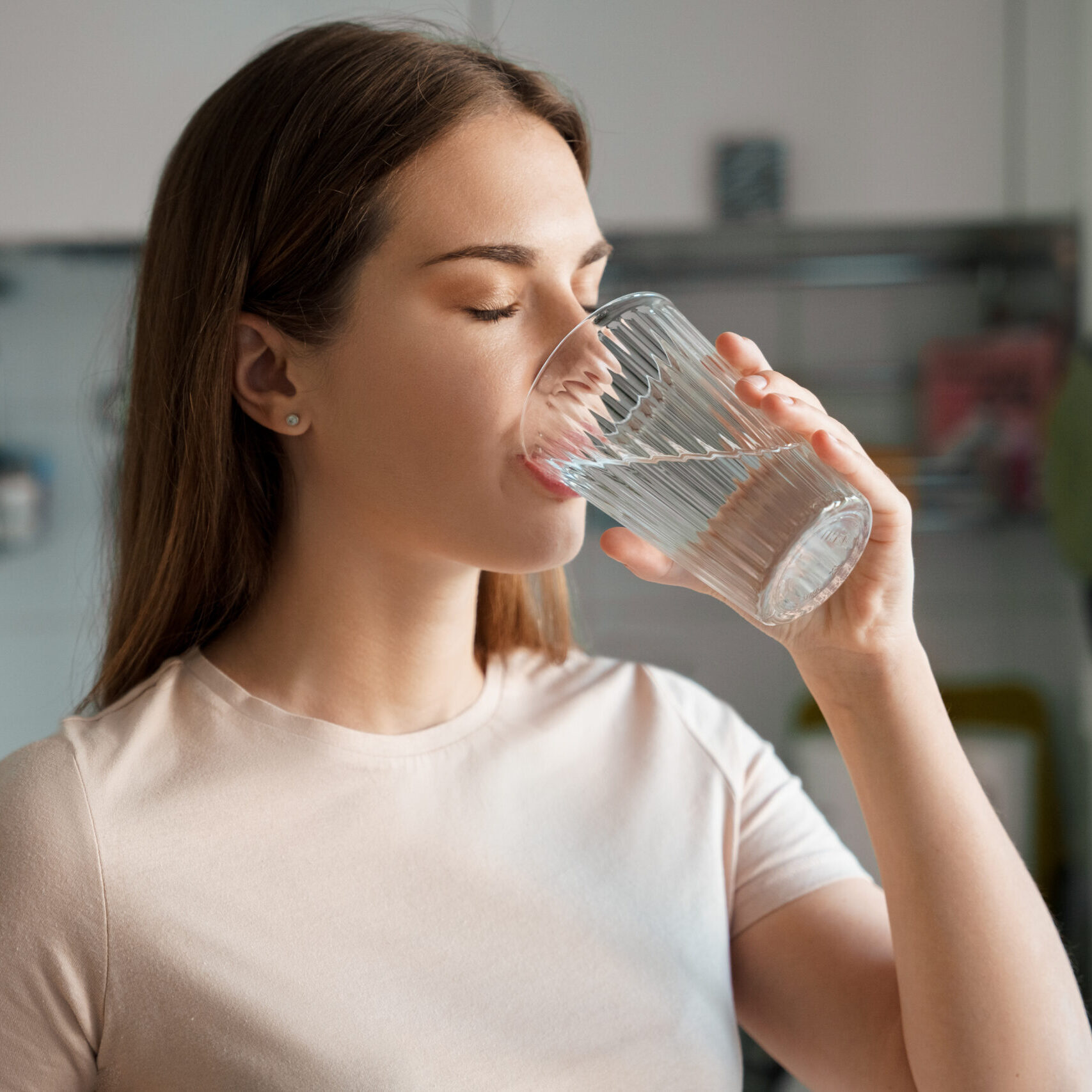 Thirsty young woman drinking fresh water from glass. Home office kitchen interior. Headshot portrait. Dehydration prevention, normal bowel function and balance of body maintenance concept