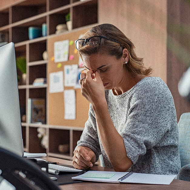 Exhausted businesswoman having a headache in modern office. Mature creative woman working at office desk with spectacles on head feeling tired. Stressed casual business woman feeling eye pain while overworking on desktop computer.
