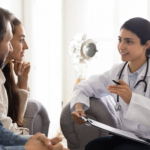 Positive young Indian doctor woman visiting couple at home, writing medical records, filling patient information file, talking to husband and wife, smiling, giving optimistic consultation